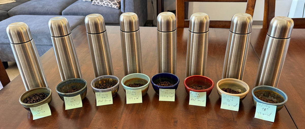 Eight insulated stainless steel travel mugs arranged in a row on a wooden table, each paired with a small ceramic bowl containing coffee.